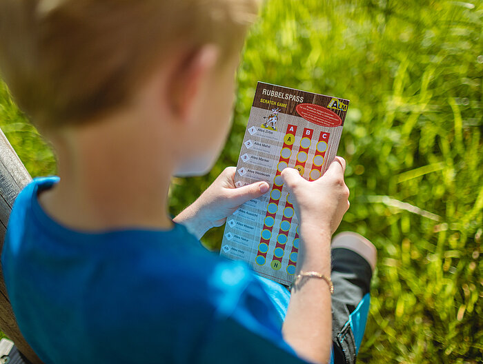 AllesAlm Ein Junge hat ein Rubbelspiel von dem Alles Alm Erlebniswegs in der Hand